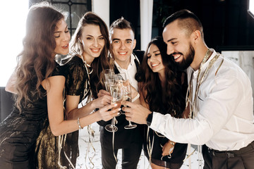 Сompany of beautiful young girls and guys dressed in stylish elegant clothes stand together and clink glasses with champagne. Party time