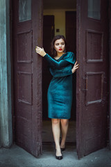 Portrait of beautiful young brunette woman with bright makeup, wearing emerald green velvet dress, stepping out the door of the old building