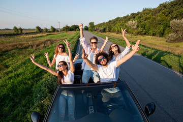 Stylish young guys are sitting and smiling in a black cabriolet on the country road on a sunny day.