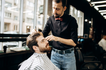 Obraz premium Barber with mustache dressed in a black shirt with a red bow tie makes a stylish hairstyle to young man in a barbershop