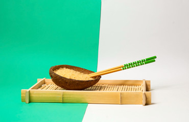 Naklejka premium Rice in organic coconut bowl with chopsticks on geometric paper background. Yellow and turquoise trendy colors, close up,copy space