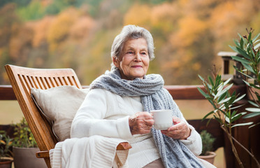 An elderly woman sitting outdoors on a terrace in on a sunny day in autumn. - obrazy, fototapety, plakaty
