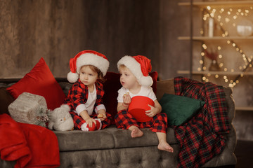 Little cute kids, brother and sister, wearing Santa Claus hats, are sitting on a big easy chair in...