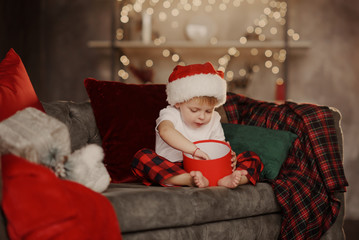A small child boy in a hat of Santa Claus sits on a soft home sofa and eats cookies from the red box. Christmas family holiday