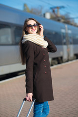 Fototapeta premium Happy young woman wearing glasses dressed in coat with big travel bag at the railway station on the train background