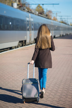 Happy young woman wearing glasses dressed in coat with big travel bag at the railway station on the train background