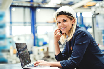 A portrait of an industrial woman engineer in a factory using laptop and smartphone.