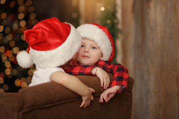 Fototapeta na wymiar Two small cute children in Santa's hats are sitting on a soft home sofa and having fun. Home Family Christmas Holiday