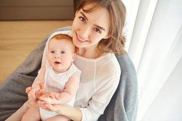 A mother with a baby in her arms sitting on a chair in front of a window in the room.