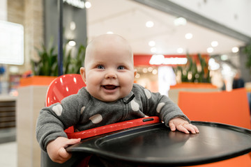 A baby in a gray suit, in a supermarket, sits in a special chair at a food court and has fun.