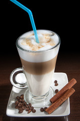 Latte macchiato in a glass with white plate, spoon, decorated with coffee beans, cinnamon. Black background.
