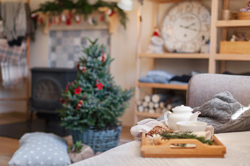 Fototapeta na wymiar Cozy home interior and decoration, tea time. Wooden serving tray on sofa near Christmas tree with red ornaments. Morning breakfast with white teapot, mug of hot beverages in living room.