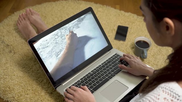 Woman looking at laptop with art sketching video on screen while sitting at home on the carpet
