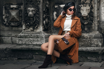 Outdoor full body fashion portrait of young fashionable woman wearing stylish glasses, beret, turtleneck, coat, ankle boots, holding suede handbag, posing in street of european city. Copy, empty space