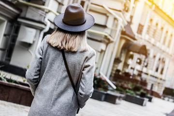 Back view of young stylish blonde woman in hat and coat walking on city streets. Attractive fashionable hipster girl standing outdoors. Modern style of city fashion. Urban outfit for young people