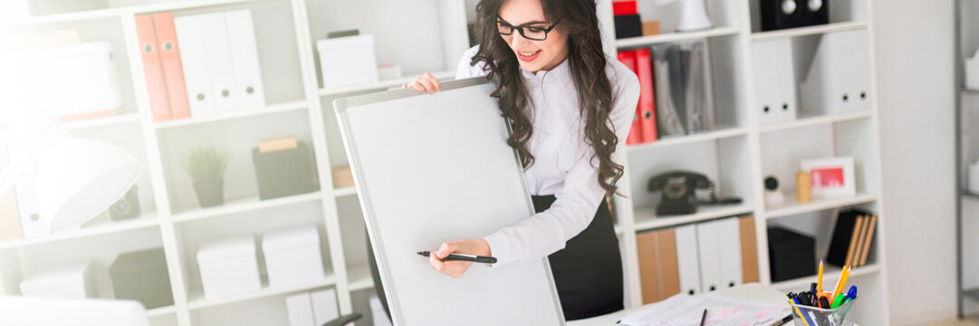 A beautiful young girl stands near an office desk and draws a magnetic marker on the magnetic board.