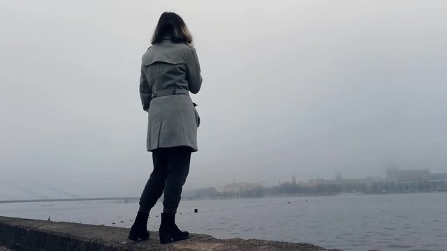 Girl standing near misty river and looking how city disappears, meanwhile vaping