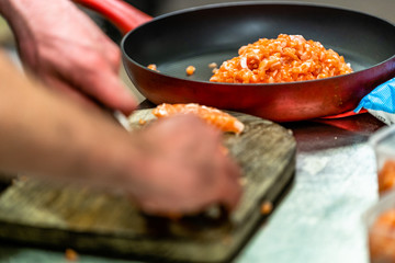 Male Chef Cutting Fresh Salmon on the Wooden Board in the Foreground with the Pan with Salmon Pieces in Focus in the Background