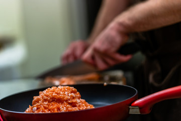 Red Pan with Pile of Cutted Salmon Pieces in the Foreground, Male Chef Cutting Fresh Salmon on the Wooden Board in the Background