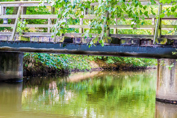 Fototapeta na wymiar Small wooden footbridge over a stream, foliage with green leaves on railing, reflection on water surface, steel beams on two concrete pillars, sunny and calm spring day in the Netherlands 