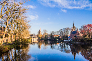Cityscape in a part of Bruges, Minnewater lake park surrounded by baretrees, buildings and medieval castle, mirror reflection on water surface, sunny winter day in province of West-Vlaanderen, Belgium