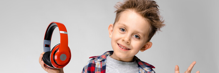 A handsome boy in a plaid shirt, gray shirt and jeans stands on a gray background. A boy in red headphones. The boy took off his headphones. © Ivan Traimak