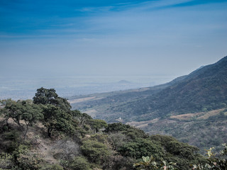 Fototapeta premium Scenic aerial view of Sierra Madre Occidental against hazy blue sky, green vegetation and trees, dry summer day at Volcanic Belt mountain range system between Jalisco and Colima states, Mexico