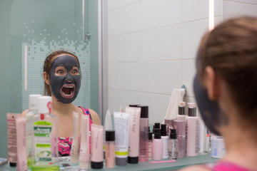 Young woman requires cosmetic mask on her face, in the bathroom and make faces at the mirror, showing different emotions