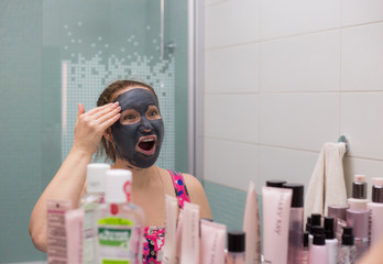 Young woman requires cosmetic mask on her face, in the bathroom and make faces at the mirror, showing different emotions