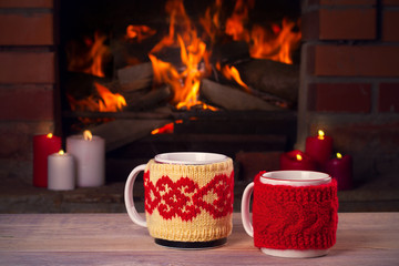 Hot drinks in mugs and candles on wooden table beside cosy open fire place. Autumn or winter holidays concept