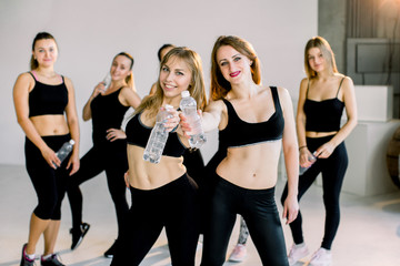 Group of young sporty girls with water boottles standing at white wall. Female companions in gym resting after fitness, indoor full length, loft studio, copy space