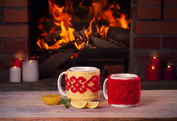 Hot drinks in mugs and candles on wooden table beside cosy open fire place. Autumn or winter holidays concept
