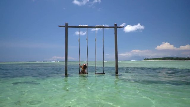 A wide shot of swing located in the crystal water with relaxing sexy brunette woman