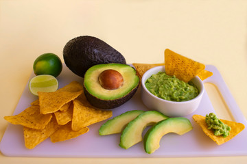 Traditional latin american mexican sauce guacamole in white bowl, avocado, lime, tortilla chips on yellow background. Copy space.