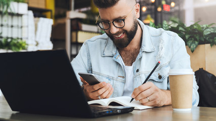 Bearded hipster man sits at table in front of laptop and writes in notebook,student prepares for exams,freelancer works.