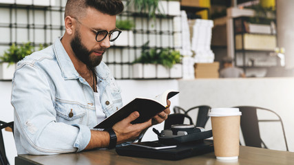 Hipster man in glasses is sitting in cafe, reading notes in notebook. On table is laptop,cup of coffee, instant camera.