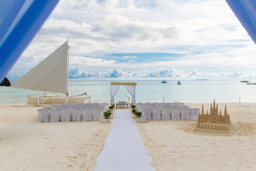 Wedding ceremony on a tropical beach in white. The arch is decorated with flowers on the sandy beach, decorated chairs for guests, sand castle and yacht in the sea. Wedding and honeymoon concept.