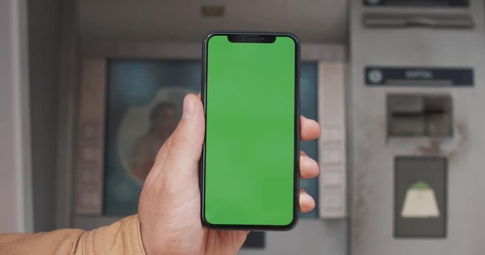 Male hands holding black smartphone with green screen on the ATM background. Big screen. Chroma key. Close up