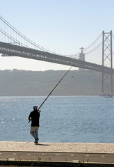 Naklejka premium A man prepares to throw out his fishing rod near Ponte 25 de Abril in Lisbon, Portugal, on a sunny day