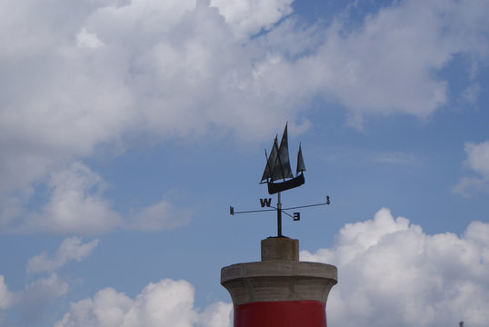 Torre roja con veleta en forma de barco velero con puntos cardinales de metal hierro sobre fondo de cielo azul y nubes blancas en d&iacute;a nublado en Puerto de Pollensa, isla de Mallorca, Islas Baleares.