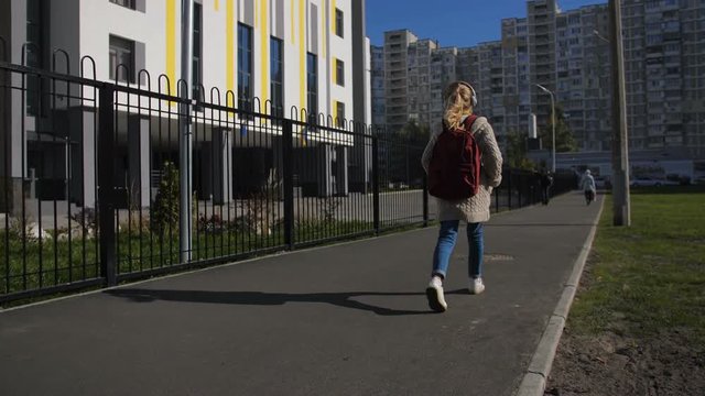 Rear view of positive blonde middle school girl with backpack walking down street, enjoying her favorite music in headphones on the way to school. Schoolgirl going to school class on sunny autumn day.