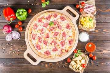 Big pizza with ham and pineapple on a round cutting board on a dark wooden background. Food ingredients.