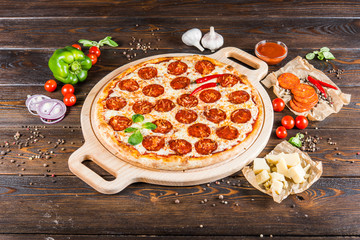 Large spicy pizza with salami and pepperoni sausage on a round cutting board on a dark wooden background. Pizza Ingredients