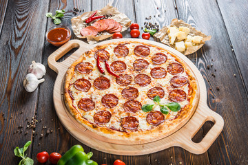 Large spicy pizza with salami and pepperoni sausage on a round cutting board on a dark wooden background. Pizza Ingredients