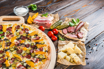 Huge pizza with sausage, pickled cucumber, bacon on a round cutting board on a dark wooden background. Pizza Ingredients