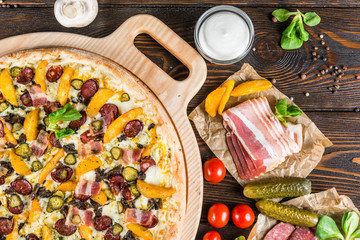 Huge pizza with sausage, pickled cucumber, bacon on a round cutting board on a dark wooden background. Pizza Ingredients