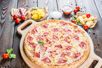 Big pizza with ham and pineapple on a round cutting board on a dark wooden background. Food ingredients. Macro
