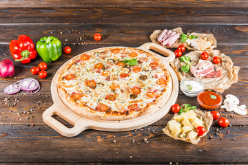 Large meat pizza with bacon, ham, chicken, sausage and tomatoes on a round cutting board on a dark wooden background. Food Ingredients.