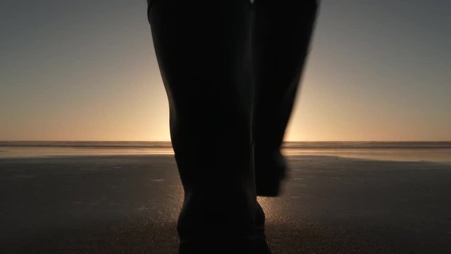 Silhouetted person walks on sandy beach towards sunset over ocean scene.