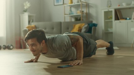 Athletic Fit Man in T-shirt and Shorts is Doing Push Up Exercises While Using a Stopwatch on His Phone. He is Training at Home in His Living Room with Minimalistic Interior. - Powered by Adobe